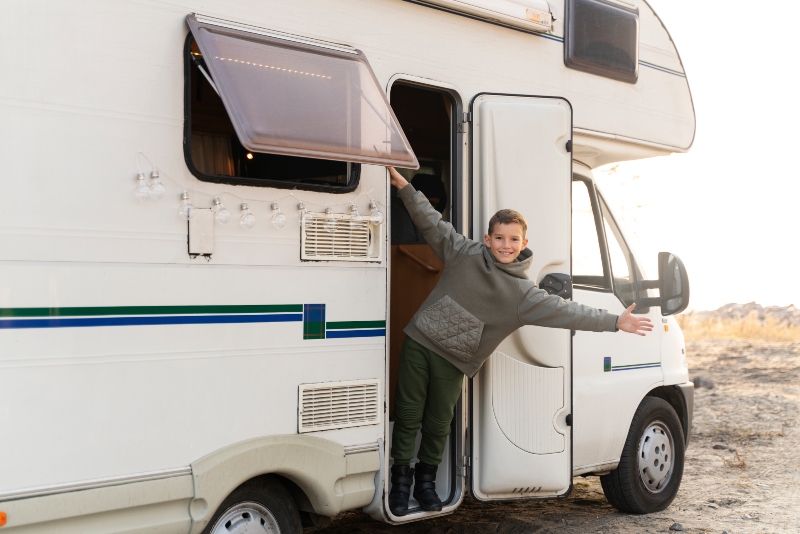 A smiling boy standing in the doorway of an RV on a sunny day, enjoying the extra space and comfort provided by the RV’s slide out system.