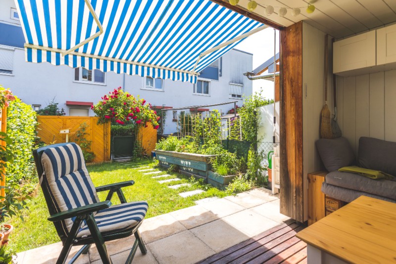 Covered patio with blue and white striped retractable awning, overlooking a cozy backyard garden with lounge seating and raised planter beds — a great example of stylish and functional patio awning ideas.