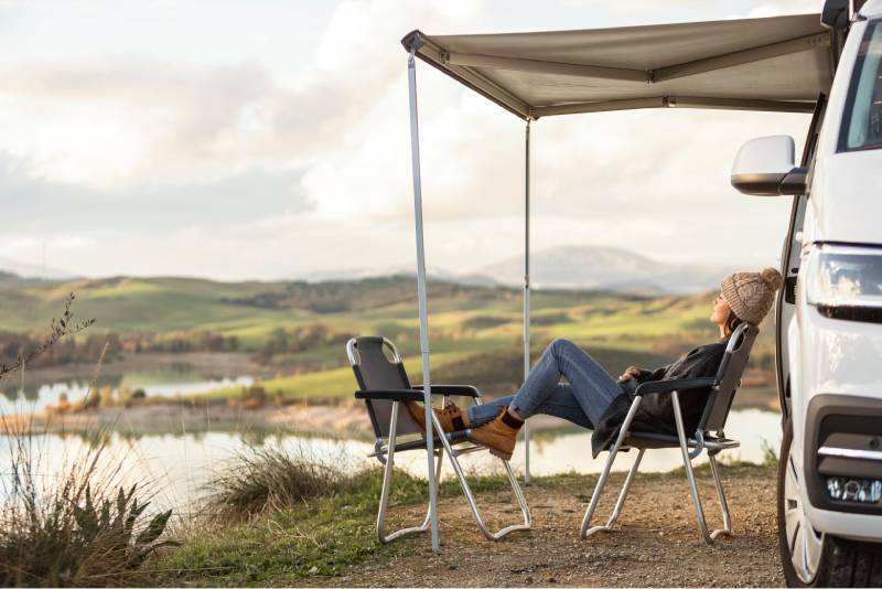A person relaxes in a camping chair under the extended awning arm of a white camper van, enjoying a scenic view of rolling green hills, a lake, and distant mountains. Dressed in a warm jacket, jeans, boots, and a knit hat, they appear content in the peaceful outdoor setting.