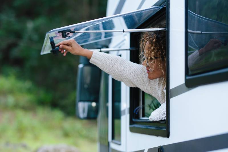 A woman reaches out through an RV window to adjust the window awning arm, possibly addressing an issue with the awning not retracting properly. The RV is parked in a natural setting with trees in the background, and she appears focused while handling the mechanism.